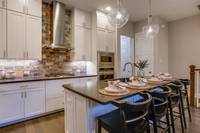 kitchen featuring a kitchen island with sink, wall chimney range hood, appliances with stainless steel finishes, decorative light fixtures, and white cabinetry