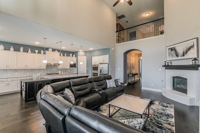 living room featuring a high ceiling, sink, a brick fireplace, ceiling fan, and dark hardwood / wood-style flooring