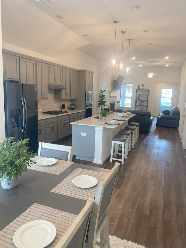 kitchen featuring backsplash, hanging light fixtures, gray cabinets, an island with sink, and refrigerator with ice dispenser