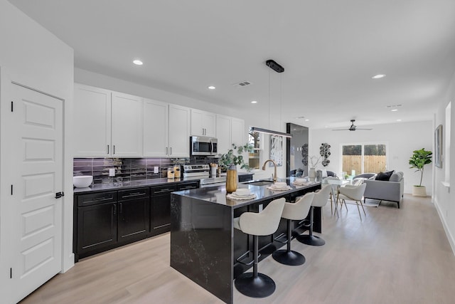 kitchen featuring stainless steel appliances, white cabinets, a center island with sink, dark countertops, and decorative light fixtures