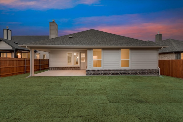 back of property at dusk with a patio, a fenced backyard, a chimney, a yard, and brick siding