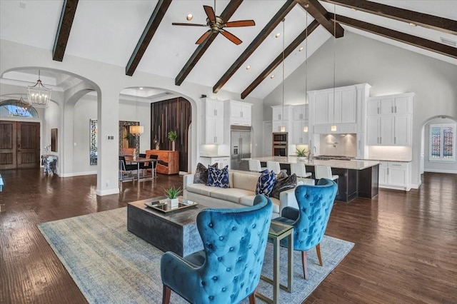 living room with beam ceiling, a towering ceiling, dark wood-type flooring, and ceiling fan with notable chandelier