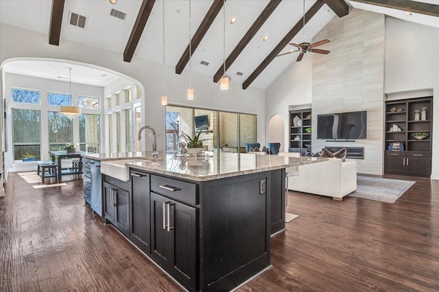 kitchen with high vaulted ceiling, a center island with sink, sink, hanging light fixtures, and light stone counters