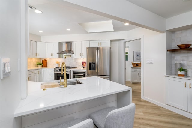 kitchen with backsplash, kitchen peninsula, wall chimney exhaust hood, appliances with stainless steel finishes, and white cabinetry