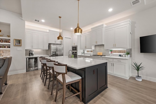 kitchen with stainless steel appliances, beverage cooler, a kitchen island with sink, decorative light fixtures, and white cabinets