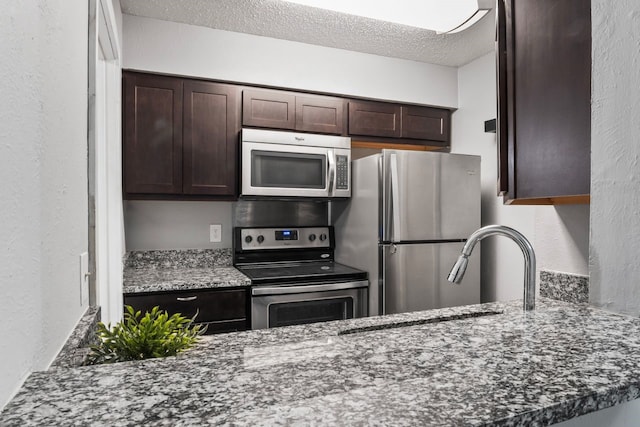 kitchen with appliances with stainless steel finishes, a textured ceiling, dark brown cabinets, and dark stone counters