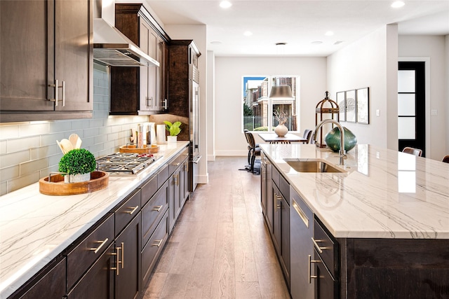 kitchen featuring sink, wall chimney range hood, an island with sink, decorative backsplash, and appliances with stainless steel finishes