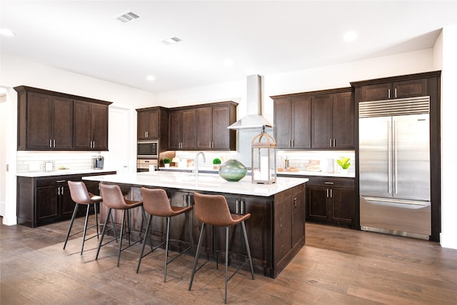 kitchen featuring dark wood-type flooring, wall chimney range hood, a kitchen breakfast bar, built in appliances, and a kitchen island with sink