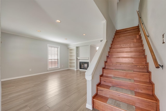 staircase with wood finished floors, a glass covered fireplace, built in features, baseboards, and recessed lighting