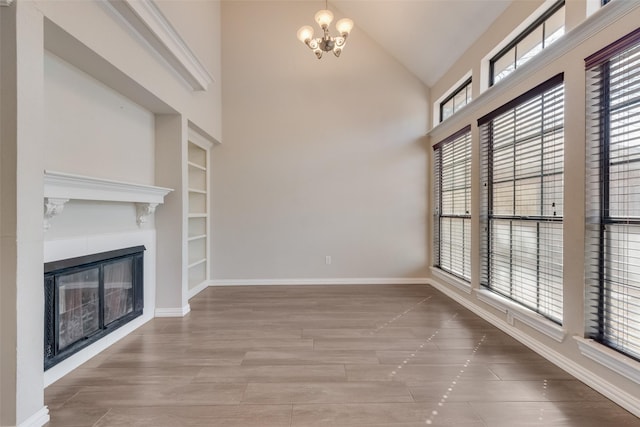 unfurnished living room with high vaulted ceiling, baseboards, a notable chandelier, a glass covered fireplace, and wood finished floors
