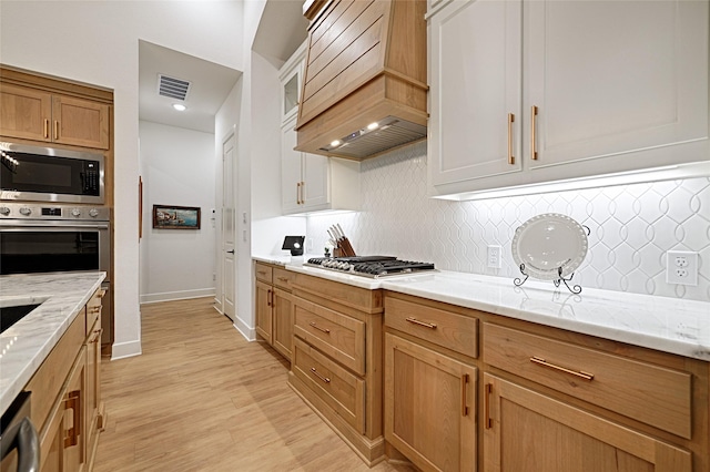 kitchen featuring light stone counters, premium range hood, light hardwood / wood-style floors, tasteful backsplash, and appliances with stainless steel finishes