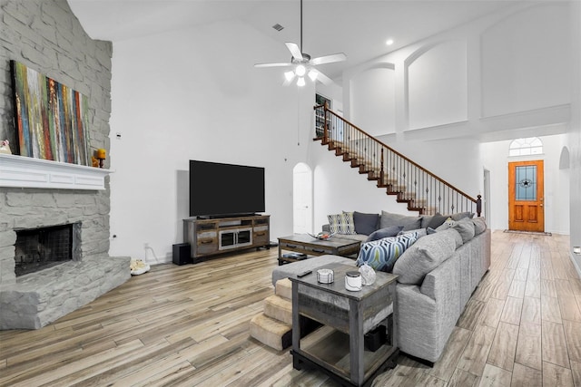 living room with ceiling fan, a stone fireplace, a towering ceiling, and light hardwood / wood-style flooring