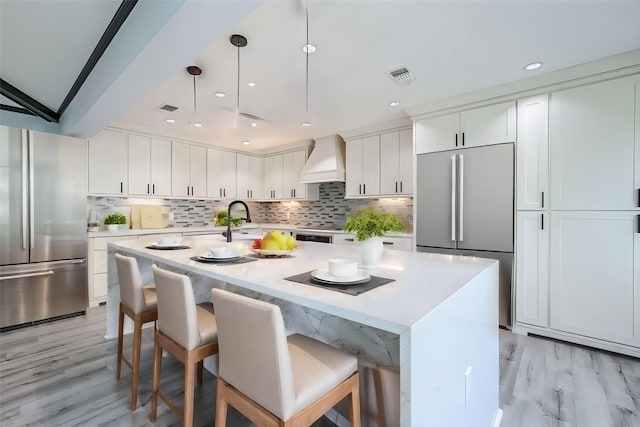 kitchen featuring premium range hood, a kitchen island with sink, white cabinets, stainless steel built in fridge, and stainless steel fridge