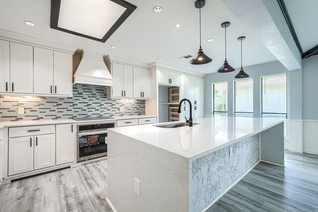 kitchen with white cabinets, sink, premium range hood, and an island with sink