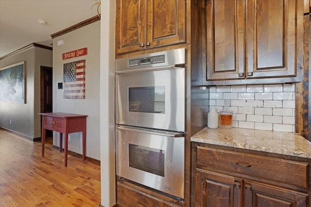 kitchen featuring decorative backsplash, ornamental molding, light stone countertops, stainless steel double oven, and light hardwood / wood-style flooring