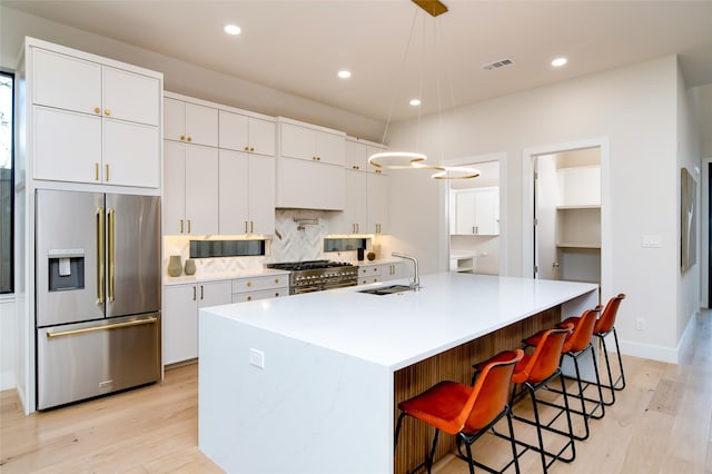 kitchen with high end appliances, a center island with sink, white cabinets, sink, and hanging light fixtures