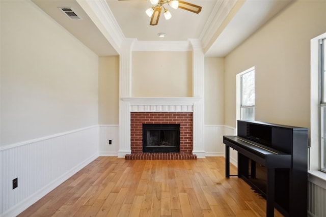 unfurnished living room featuring light wood-style flooring, a wainscoted wall, visible vents, a brick fireplace, and crown molding