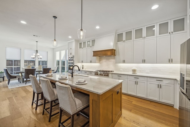 kitchen with custom exhaust hood, white cabinets, light stone countertops, an island with sink, and decorative light fixtures