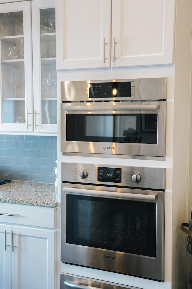 kitchen featuring stainless steel double oven, backsplash, white cabinetry, and light stone counters