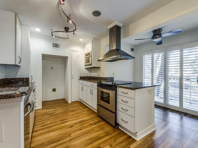 kitchen with light wood-type flooring, ceiling fan, wall chimney range hood, electric range, and white cabinetry