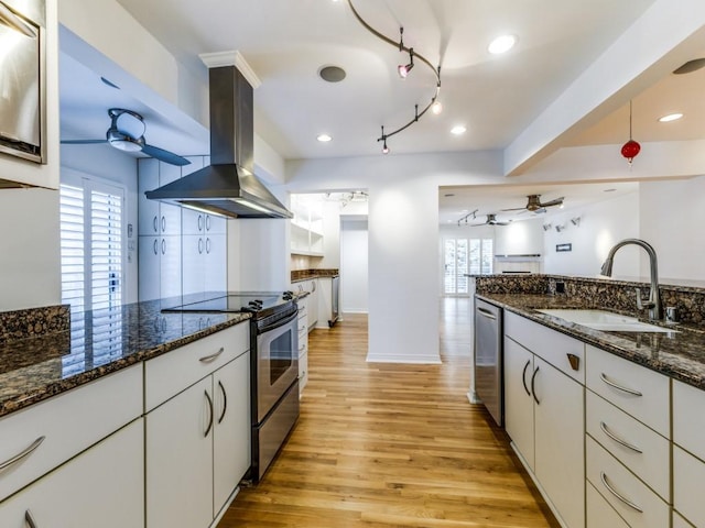 kitchen featuring wall chimney exhaust hood, stainless steel appliances, ceiling fan, sink, and white cabinetry