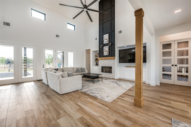 living room featuring ceiling fan, french doors, a stone fireplace, a towering ceiling, and light wood-type flooring