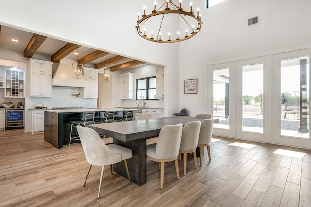 dining space featuring an inviting chandelier, a towering ceiling, beamed ceiling, light hardwood / wood-style floors, and beverage cooler