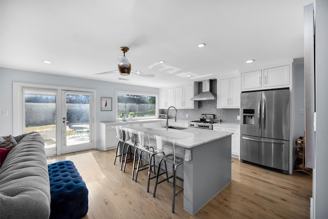 kitchen with wall chimney range hood, stainless steel refrigerator with ice dispenser, sink, white cabinetry, and a kitchen island with sink