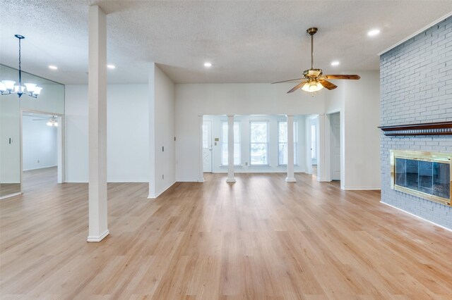 unfurnished living room with ornate columns, a brick fireplace, a textured ceiling, ceiling fan with notable chandelier, and light wood-type flooring