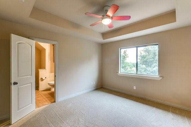 unfurnished bedroom with ceiling fan, ensuite bath, a tray ceiling, and light carpet