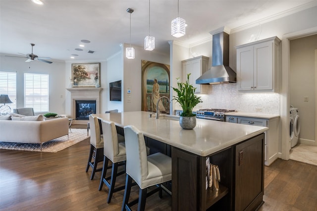 kitchen featuring wall chimney exhaust hood, decorative light fixtures, stainless steel range, independent washer and dryer, and a kitchen island with sink