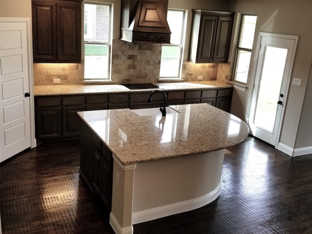 kitchen featuring light stone counters, custom exhaust hood, sink, a center island with sink, and dark brown cabinetry