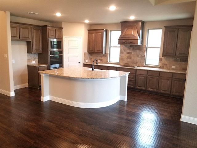 kitchen with stainless steel appliances, sink, light stone counters, premium range hood, and a center island with sink