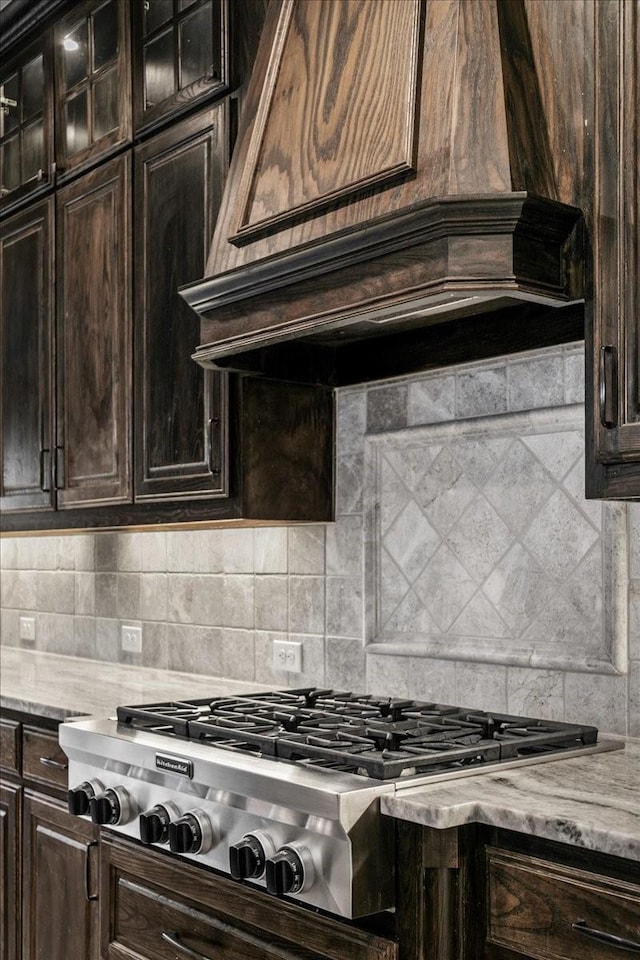 kitchen with stainless steel gas stovetop, decorative backsplash, light stone countertops, custom range hood, and dark brown cabinetry