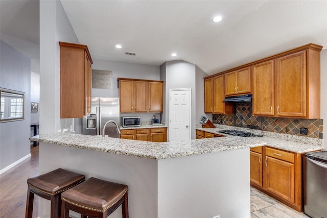 kitchen featuring ventilation hood, appliances with stainless steel finishes, a kitchen breakfast bar, and light stone counters