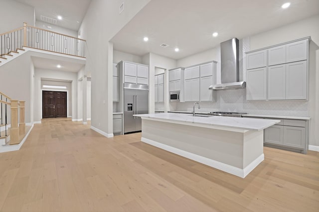 kitchen with a kitchen island with sink, wall chimney range hood, sink, built in appliances, and light hardwood / wood-style floors