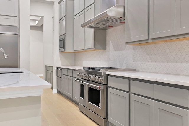 kitchen with ventilation hood, built in appliances, gray cabinets, light wood-type flooring, and light stone counters