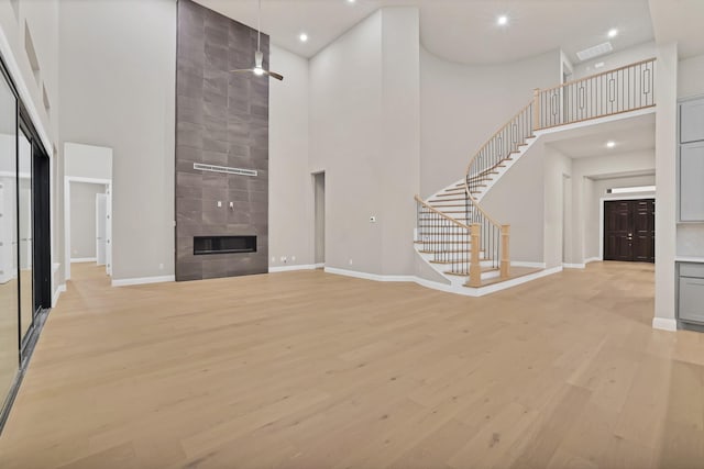 unfurnished living room with ceiling fan, a towering ceiling, a tile fireplace, and light hardwood / wood-style flooring