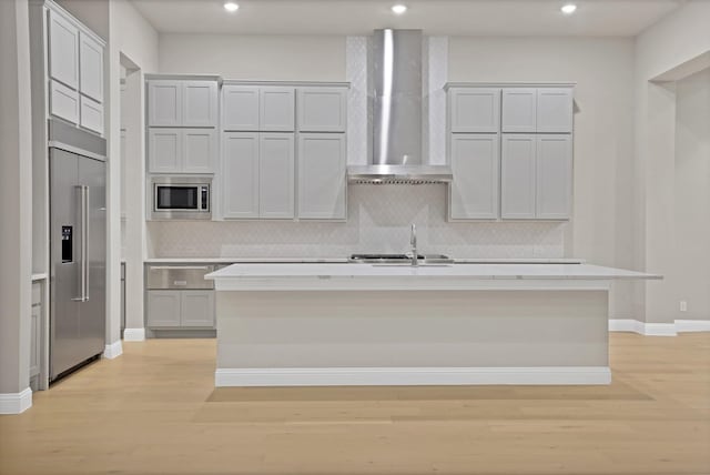 kitchen with backsplash, a center island with sink, sink, built in appliances, and wall chimney exhaust hood