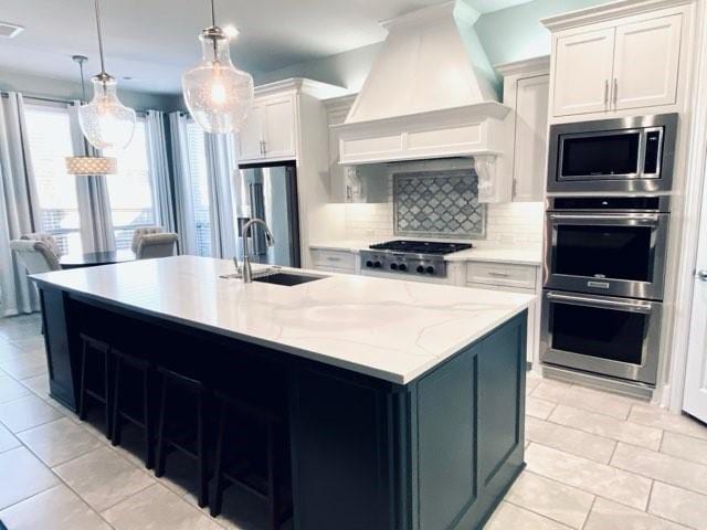 kitchen featuring tasteful backsplash, custom range hood, stainless steel appliances, decorative light fixtures, and white cabinetry