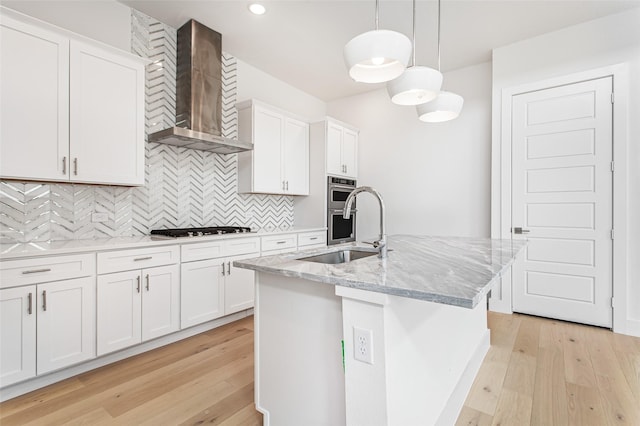 kitchen featuring a kitchen island with sink, sink, wall chimney range hood, decorative light fixtures, and white cabinetry