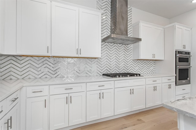 kitchen with wall chimney range hood, decorative backsplash, light stone countertops, white cabinetry, and stainless steel appliances