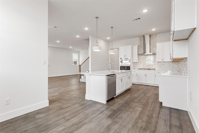 kitchen with wall chimney exhaust hood, hanging light fixtures, an island with sink, stainless steel appliances, and white cabinets