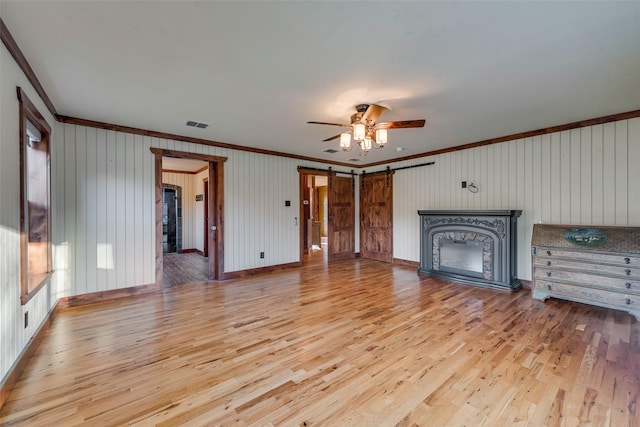 unfurnished living room with crown molding, ceiling fan, a barn door, and light hardwood / wood-style floors