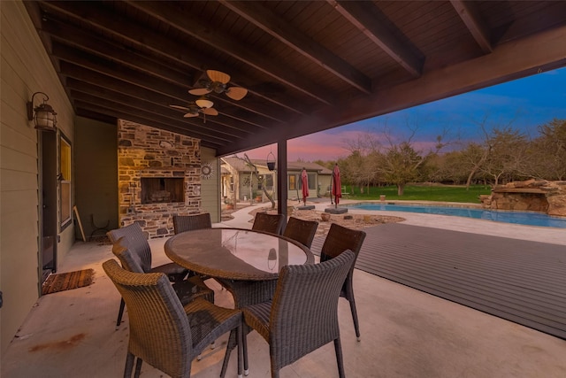 patio terrace at dusk featuring ceiling fan and an outdoor stone fireplace