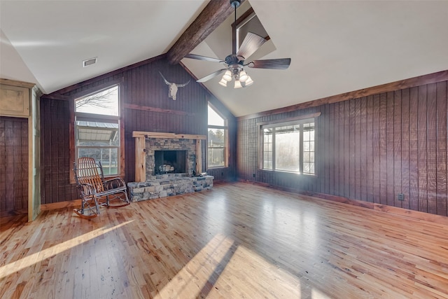 unfurnished living room featuring a stone fireplace, wood walls, light hardwood / wood-style flooring, ceiling fan, and beam ceiling