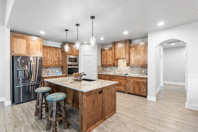 kitchen with tasteful backsplash, light stone counters, stainless steel appliances, a kitchen island with sink, and hanging light fixtures