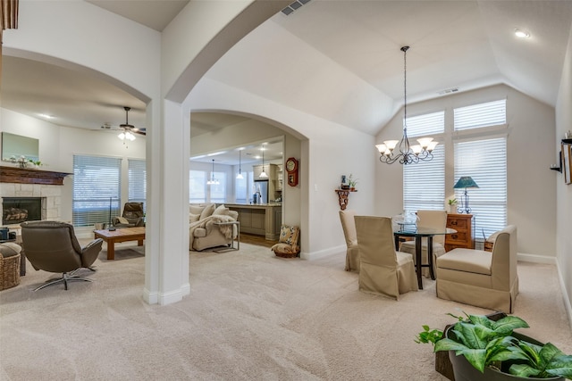 carpeted dining area featuring ceiling fan with notable chandelier, a stone fireplace, and vaulted ceiling