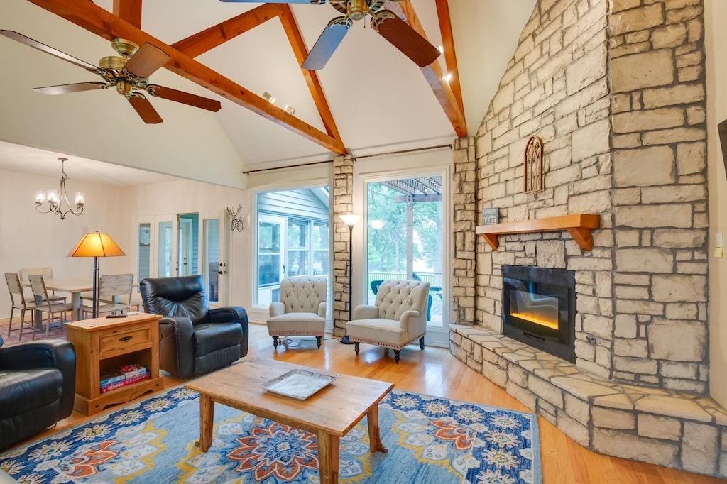 living room with a stone fireplace, beamed ceiling, ceiling fan with notable chandelier, and light wood-type flooring