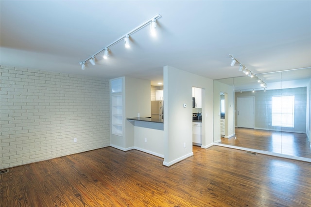 unfurnished living room with dark hardwood / wood-style flooring, track lighting, and brick wall